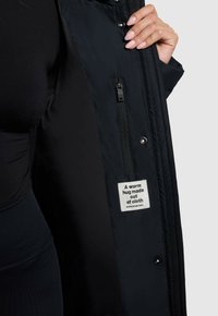 Zwarte buitenjack met een ritsvak aan de binnenzijde, met een label waarop staat "Een warme omhelzing van stof." Gladde stoffen textuur.