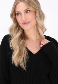 Schwarzer, strukturierter Pullover mit V-Ausschnitt und langen Ärmeln. Modell mit welligem blonden Haar und natürlichem Make-up hält einen Teil des Pullovers.