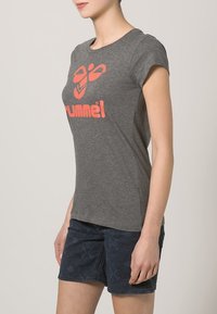 Camiseta gris de manga corta con un gran logotipo gráfico naranja en el frente. Tejido suave, forma ajustada, diseño minimalista. Combinada con pantalones cortos oscuros con un estampado.
