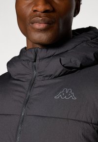 Giacca nera isolante con chiusura a zip e cappuccio. Presenta una superficie testurizzata e un logo discreto sul petto. Materiale liscio.