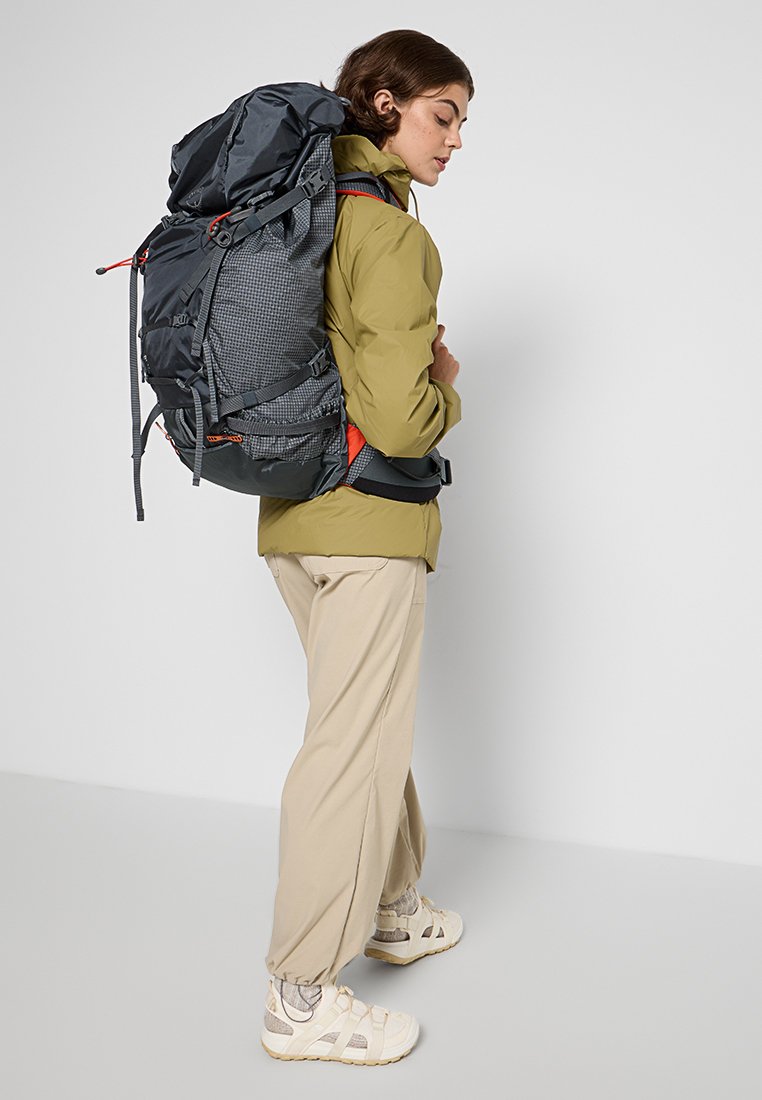 Rugzak van donkergrijs stof met een gestructureerd patroon, verstelbare banden en feloranje accenten. Model draagt een olijfgroen jack en beige broek.