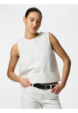 Jeune femme aux cheveux bruns attachés porte un haut blanc sans manches et un pantalon blanc avec une ceinture noire, debout les mains dans les poches.