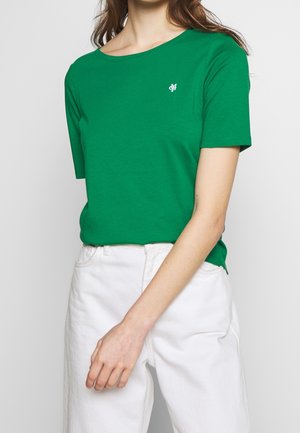 Kobieta ubrana w zielony t-shirt z krótkim rękawem z małym białym logo na piersi, dobrany do białych spodni z wysokim stanem.