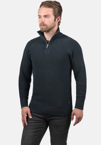 Suéter de punto azul marino con cuello con cremallera, puños y dobladillo acanalados, que presenta un patrón texturizado y un acento de logo en la parte inferior delantera.