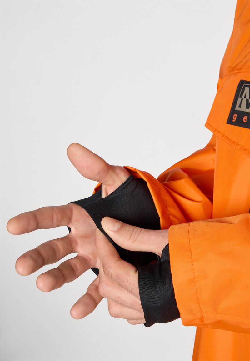Giacca impermeabile di un vivace arancione con polsini neri. Presenta aperture per il pollice per l'esposizione delle mani. Tessuto morbido e leggero con una vestibilità rilassata.