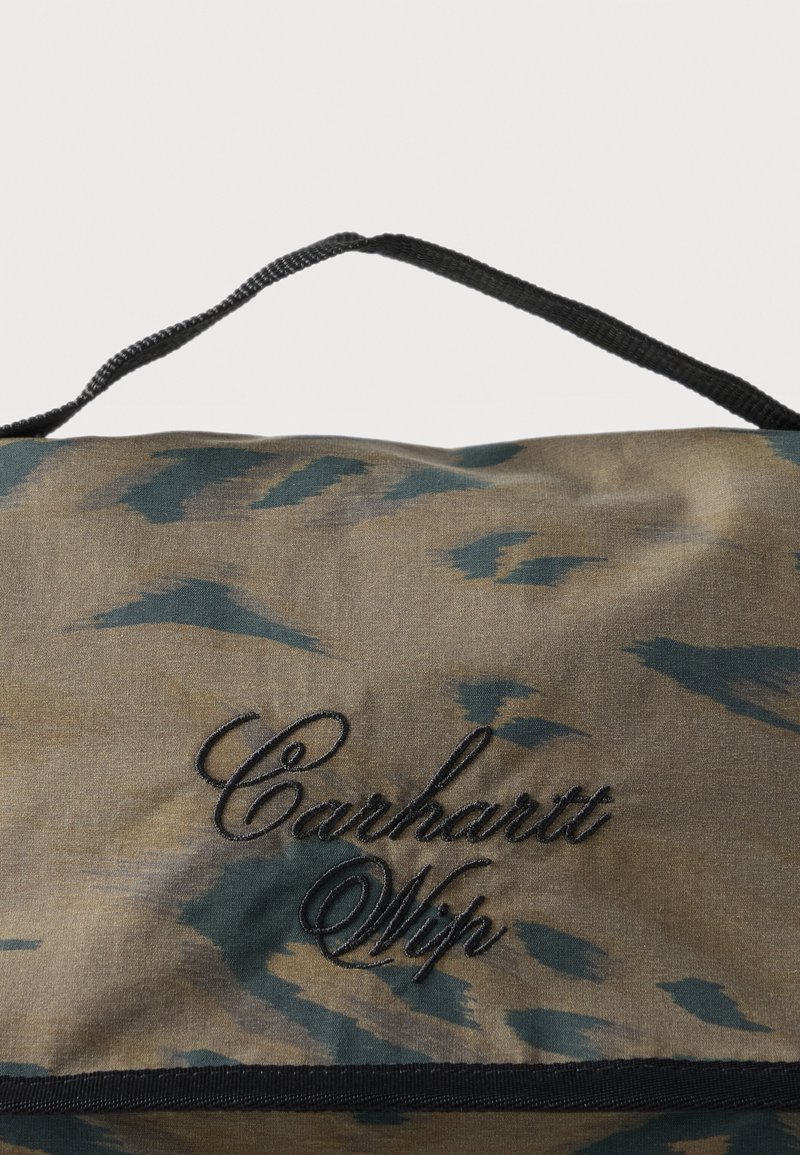 Bolsa de lona con patrón de camuflaje verde y marrón y texto bordado en negro "Carhartt Wip", con asa de tela negra.