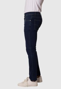 Jeans de mezclilla azul oscuro con corte ajustado, bolsillos estándar y costuras amarillas en contraste, combinados con zapatillas blancas.