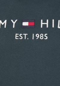 Tejido texturizado verde oscuro con texto bordado en blanco "TOMMY HILFIGER EST. 1985" y un pequeño logo de bandera en azul, blanco y rojo.