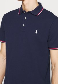 Tmavě modré polo tričko z bavlny s texturovaným povrchem. Má tříknoflíkovou légu, bílý logo na hrudi a manžety s červeno-bílými pruhy.