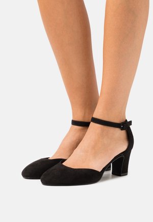 Chaussures à talons moyens en suède noir avec des brides à la cheville, portées sur des jambes de teinte claire, présentées contre un fond blanc uni.