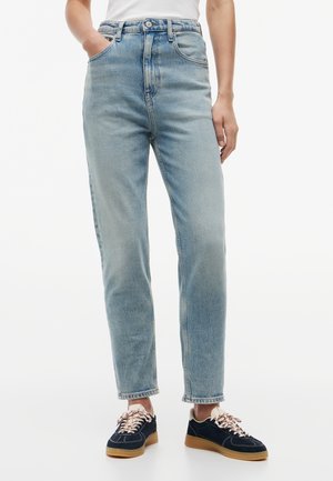 Persoană purtând blugi albaştri deschis, cu talie înaltă și tăietură dreaptă, cu adidași bleumarin prevăzuți cu șireturi albe și roșii, stând în picioare pe un fundal alb.