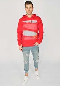 Rød hættetrøje med et grafisk design, der har grå striber og gule accenter, lavet af bomuld. Bæres med slidte lyseblå jeans og hvide sneakers.