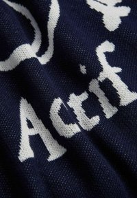 Navy knitted textile featuring white text, displaying letters in a bold, modern font with visible texture and subtle pattern details.
