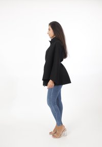 Svart kappa med hög krage, har en utsvängd nederdel, tillsammans med blå tajta jeans och transparenta klacksandaler.