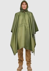 Vihreä vedenpitävä poncho hupulla, tekstuuri pinnassa ja napsautettavat sulkimet sivuilla. Muotoiltu käytettäväksi beigen housujen ja saappaiden päällä.