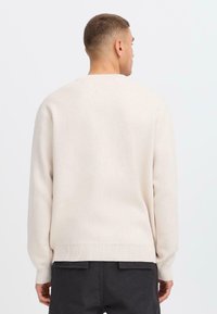 Lichtbeige trui, met een gestructureerde brei, ronde hals, ribbels bij de mouwen en onderkant, relaxed fit, gedragen met donkergrijze broek, gezien van achter.