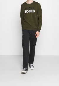 Felpa verde oliva con il testo "JACK&JONES" in bianco e nero. Abbinata a pantaloni cargo neri e sneakers nere, con lacci bianchi.