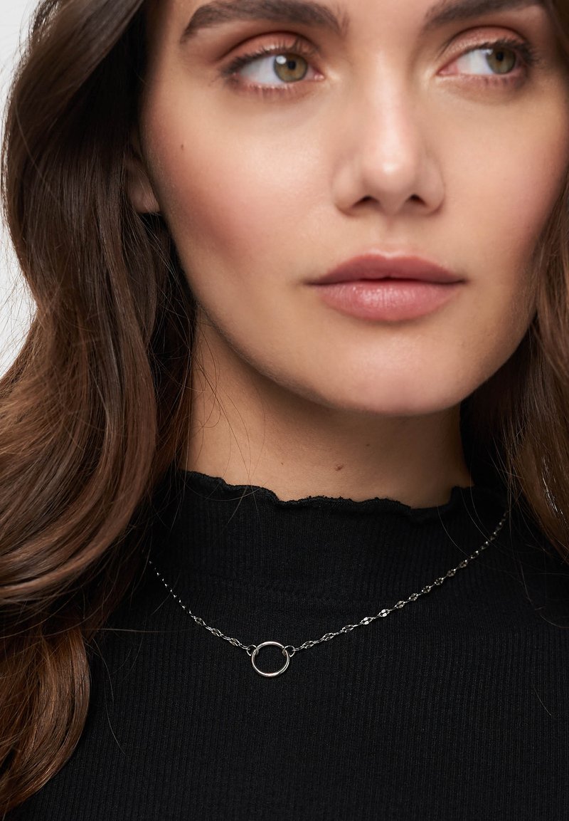 Silberne Halskette mit einem runden Anhänger, getragen von einer Person in einem schwarzen, gerippten Rollkragenpullover mit gewelltem Saum. Das Haar ist lang und braun.