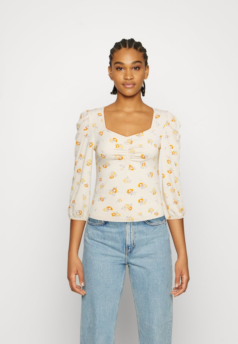 Top floral bege claro com mangas bufantes, decote quadrado e detalhes franzidos na frente; apresenta padrões florais amarelos e laranjas.