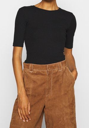 Kobieta w dopasowanym czarnym prążkowanym topie z rękawami do łokcia oraz wysokim stanem brązowych zamszowych spodniach, z jedną ręką w kieszeni.