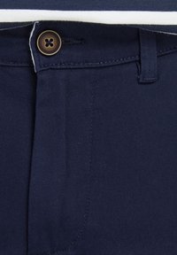 Gros plan d'un pantalon bleu marine avec un bouton marron, une boucle de ceinture et des coutures visibles près de la taille et de la braguette.