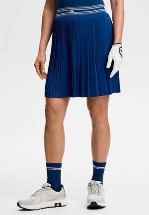 Person iført en plisseret navyblå nederdel, matchende navy strømper med hvide striber, hvide sneakers og en hvid golfhandske på højre hånd.