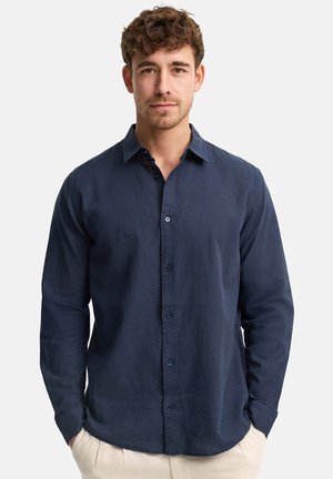 Jeune homme aux cheveux courts et bouclés, portant une chemise bleu foncé boutonnée et un pantalon clair, debout les mains dans les poches.