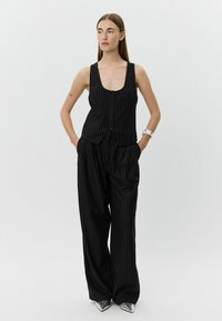 Femeie îmbrăcată cu un veston negru cu dungi și pantaloni evazați, încălțată cu pantofi argintii ascuțiți, stând cu mâinile în buzunare pe un fundal simplu.