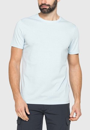 Uomo con barba che indossa una maglietta azzurro chiaro a maniche corte e pantaloni cargo scuri, in piedi davanti a uno sfondo chiaro e uniforme.