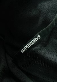 Melnas vilnas audums ar teksturētu virsmu. Iespējams, izceļas izteikts balts "SUPERDRY" logotips uz melna joslas, demonstrējot kontrastu un zīmola identitāti.