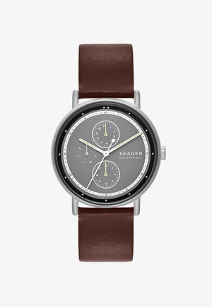 Pyöreä hopeinen kello, jossa on harmaa kellotaulu, kaksi alinäyttöä, vihreät korostukset ja ruskea nahkaranneke. Kello on minimalistinen muotoilultaan ja sen merkit ovat selkeitä.