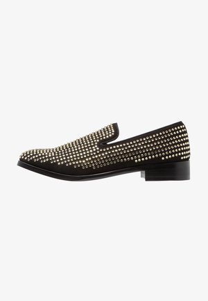 Chaussure noire à enfiler avec un petit talon, recouverte de petits clous dorés ronds, espacés uniformément, vue de côté extérieur.