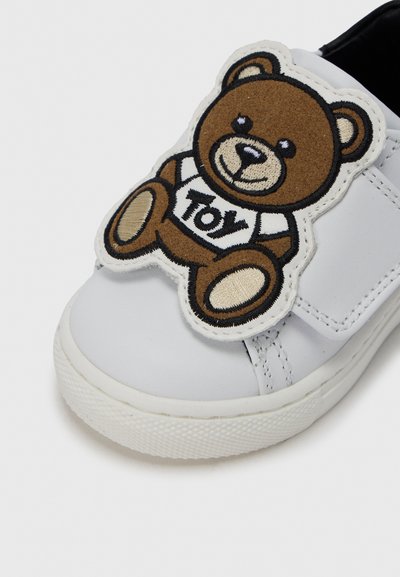 Λευκό συνθετικό αθλητικό παπούτσι με μεγάλο καφέ μπαλώμα αρκουδάκι που φοράει μπλουζάκι με τη λέξη "Toy", λουράκι με βέλκρο και υφασμένο λαστιχένιο πέλμα.