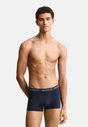 Junger Mann mit kurzen lockigen Haaren, der marineblaue GANT Boxershorts trägt, steht vor einem schlichten hellgrauen Hintergrund.