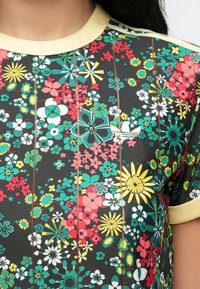 Κοντινή λήψη πολύχρωμου πουκαμίσου με λουλουδένιο μοτίβο, κίτρινο ribbed γιακά και μανίκια, με μικρό λευκό λογότυπο Adidas στο στήθος.