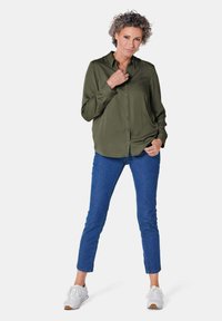 Olivgrünes Hemd mit Knopfleiste und langen Ärmeln, blaue, enge Jeans und weiße Sneakers. Glatte Textur, klassischer Kragen und lässige Passform.
