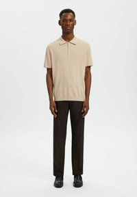 Homme debout portant un polo beige à manches courtes avec col zippé, un pantalon droit foncé et des mocassins noirs sur fond blanc uni.