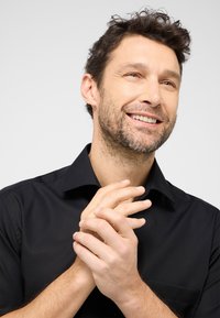 Zwart overhemd met knopen en een kraag, gladde textuur en een borstzak. Model heeft kort, krullend haar en klapt handen.