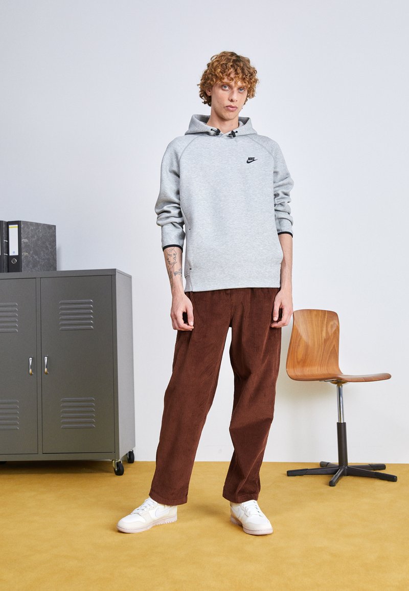 Hoodie gris avec logo noir, associé à un pantalon en velours côtelé marron et à des baskets blanches. Design simple avec une texture côtelée, coupe décontractée.