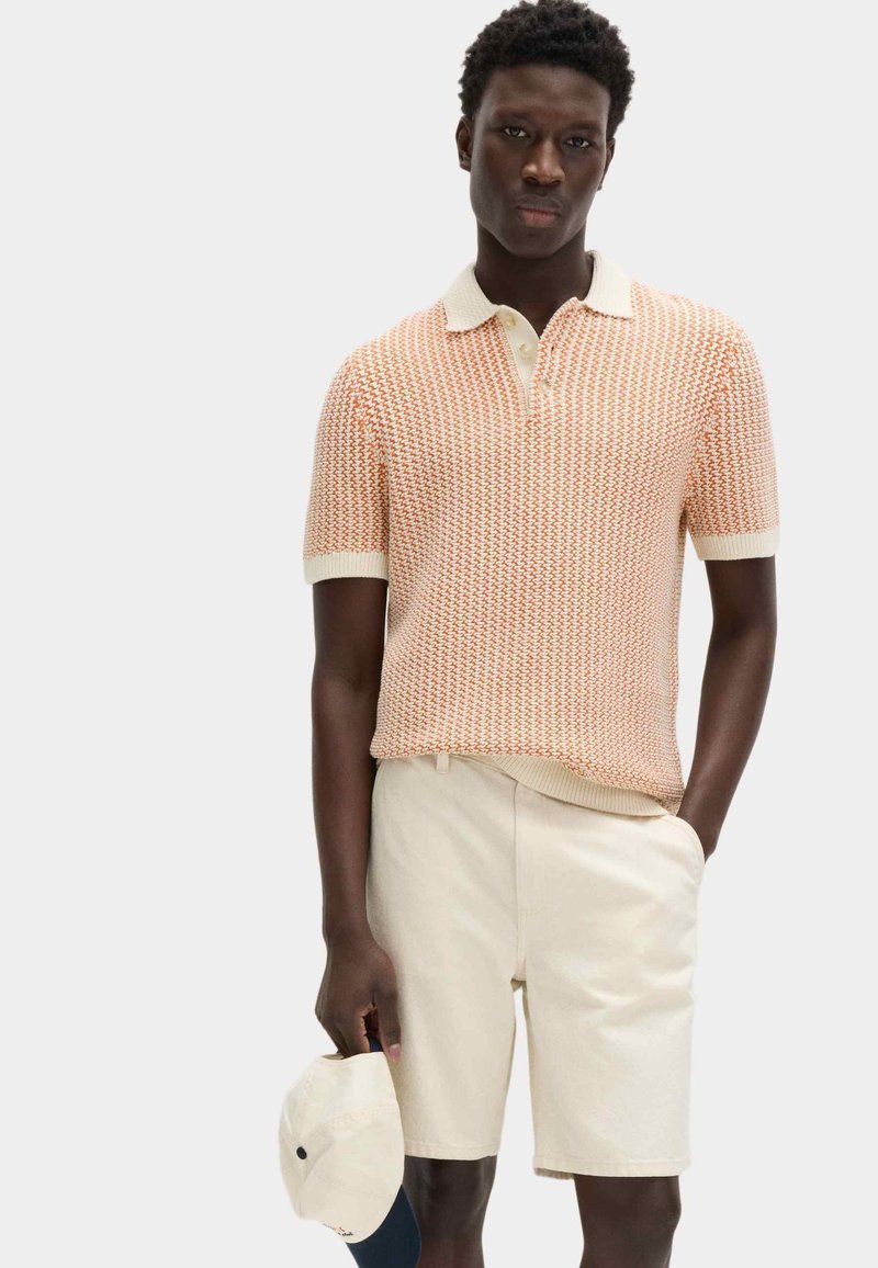 Ung man som bär en kortärmad orangemönstrad pikétröja och crèmefärgade shorts, håller en vit basebollkeps vid sin sida.