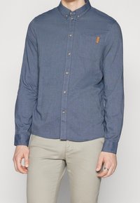 Camicia a maniche lunghe con bottoni in tessuto blu scuro, con tasca sul petto e un accento in pelle. Tessuto liscio con cuciture leggere.