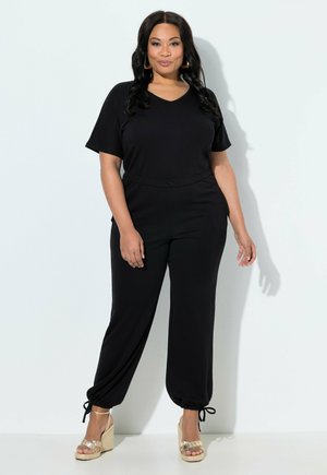 Donna in piedi contro un muro bianco, indossa una maglietta nera a maniche corte, pantaloni neri con laccetti alla caviglia e sandali zeppa dorati.