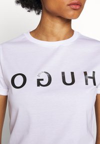 Camiseta de algodón blanca con un destacado texto negro invertido que dice "HUGO" en el pecho. Textura suave con un clásico escote redondo.