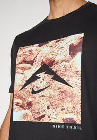 Sort t-skjorte med et fjellmotiv og Nike-logo i svart over en teksturert steinbakgrunn med teksten "Nike Trail" i lyseblått.