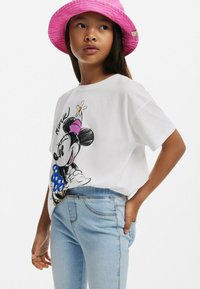 Bílá bavlněná tričko s barevným grafickým motivem Minnie Mouse. V kombinaci se světlými modrými džíny. Modelka má na sobě jasně růžovou kšiltovku.