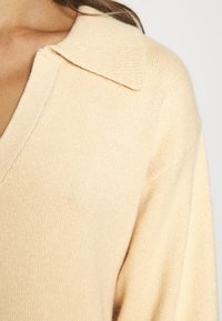 Ljusbeige stickad tröja med V-ringad hals och stor krage. Tyget har en mjuk, något strukturerad yta.