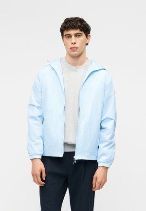 Jeune homme portant une veste à capuche bleu clair par-dessus un sweat-shirt gris et un pantalon noir, debout devant un fond blanc uni.