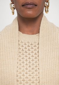 Suurendatud lähedus inimesest, kes kannab paksu kuldset rõngaskõrvarõngast ja kreemivärvi tekstureeritud kudumit koos sobiva salliga.