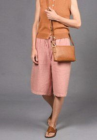 Femme portant un haut sans manches marron, un short rose ample jusqu'aux genoux, des sandales à lanières marron et un petit sac bandoulière marron, se tenant sur un fond gris.