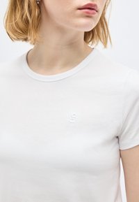 Γυναίκα που φοράει ένα απλό λευκό T-shirt με στρογγυλή λαιμόκοψη και μικρό ανάγλυφο λογότυπο αριστερά στο στήθος, ορατή από τους ώμους έως το μέσο του κορμού.